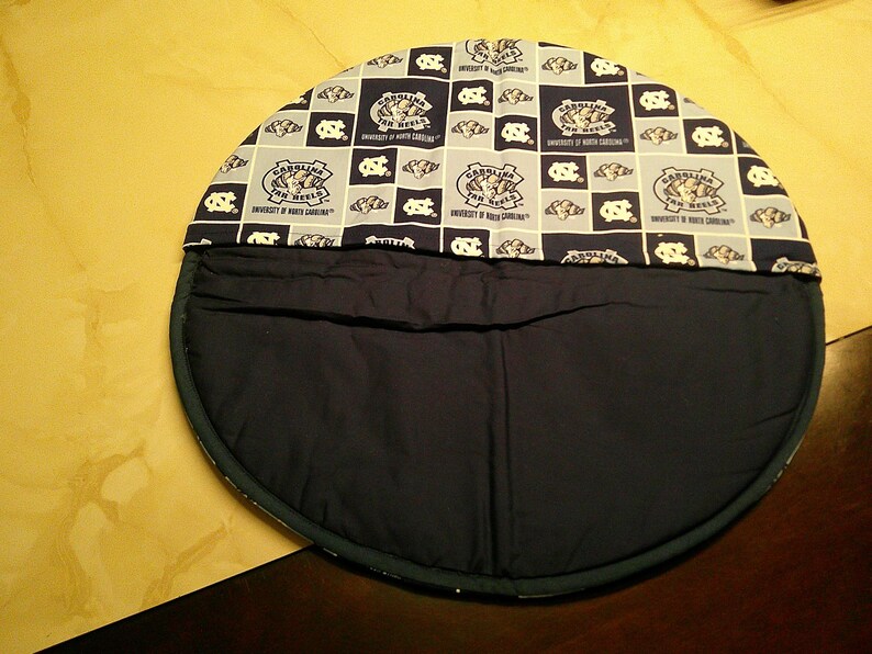May include: A round fabric tortilla warmer featuring a navy blue and light blue checkered pattern with the University of North Carolina Tar Heels logo and text printed repeatedly. The bottom half is solid navy blue fabric with a circular stitched edge.