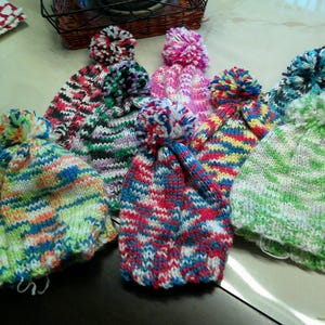 May include: A collection of colorful, knitted beanies with pom-poms. The hats feature various color combinations, including green, blue, red, and pink, with white accents. The pom-poms are also multi-colored.