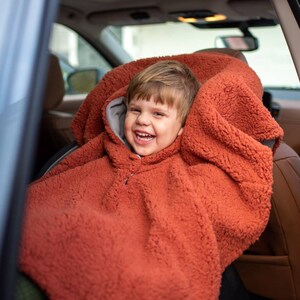 Op de afbeelding: Een lachend kind draagt een roestkleurige, met capuchon bedekte autostoel. De hoes is gemaakt van een zacht, fuzzy materiaal met een grijze voering rond de capuchon. Het kind zit in een auto met bruine leren stoelen.