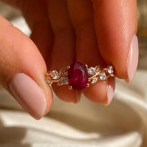 May include: A gold ring featuring a central oval-cut deep red ruby gemstone. The band is designed with intricate metal leaf motifs and small, round, clear white accent stones embedded along the sides of the setting, creating a botanical look.
