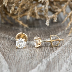 May include: A pair of gold stud earrings with round, clear gemstones. The earrings have a screw-back closure.