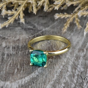 Op de afbeelding: Een gouden ring met een smaragdgroene edelsteen in kussenslijp. De ring ligt op een getextureerd grijs oppervlak.