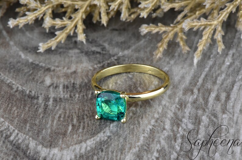 Op de afbeelding: Een gouden ring met een smaragdgroene edelsteen in kussenslijp. De ring ligt op een getextureerd grijs oppervlak.