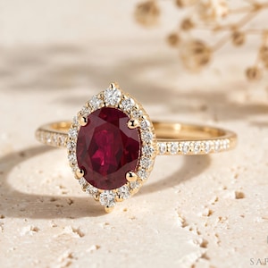 May include: A gold ring featuring a large oval-cut deep red ruby center stone surrounded by a halo of small round white diamonds. The band is also set with small white diamonds along the sides. The ring is marked with the Sapheena logo in the corner.