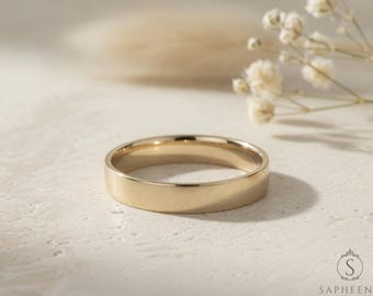 May include: A gold-colored wedding band with a flat profile. The ring is centered on a textured, off-white surface, with soft lighting and a blurred background. Delicate white flowers and foliage are in the background.