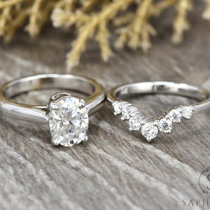 May include: A set of two silver engagement rings with a large oval-shaped diamond on the main ring and a smaller diamond band with a curved design.