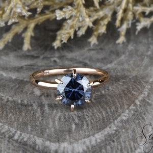 Puede incluir: Un anillo de compromiso de oro rosa con un engaste de seis garras y una gran piedra preciosa redonda de color azul oscuro.