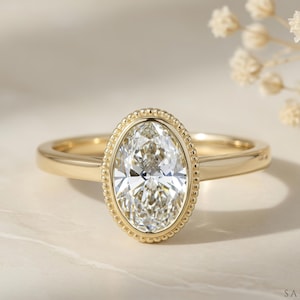 May include: A gold ring with an oval-cut diamond center stone. The diamond is surrounded by a beaded halo. The band is smooth and polished. The ring is set against a neutral background with a sprig of white flowers.