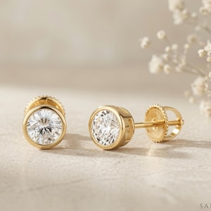 May include: A pair of gold stud earrings featuring round, clear, faceted stones set in a bezel setting. Each earring includes a threaded post and a matching gold screw-back closure. The design is simple, with a polished yellow gold finish.