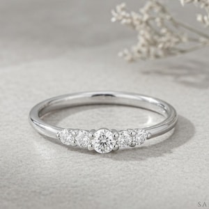 Peut inclure: Une bague en métal argenté ornée d'une pierre précieuse transparente ronde centrale, flanquée de deux pierres plus petites de chaque côté, totalisant cinq pierres sur le devant de l'anneau fin et poli, avec la marque Sapheena visible.