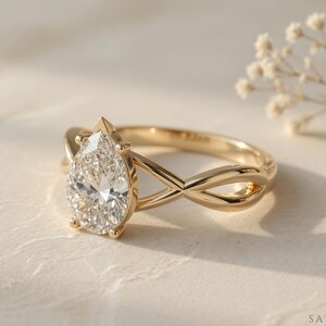 May include: A gold engagement ring with a pear-cut diamond. The ring has an infinity-style band. The diamond is the focal point, sparkling brightly. The ring is set against a neutral background, with delicate floral accents.