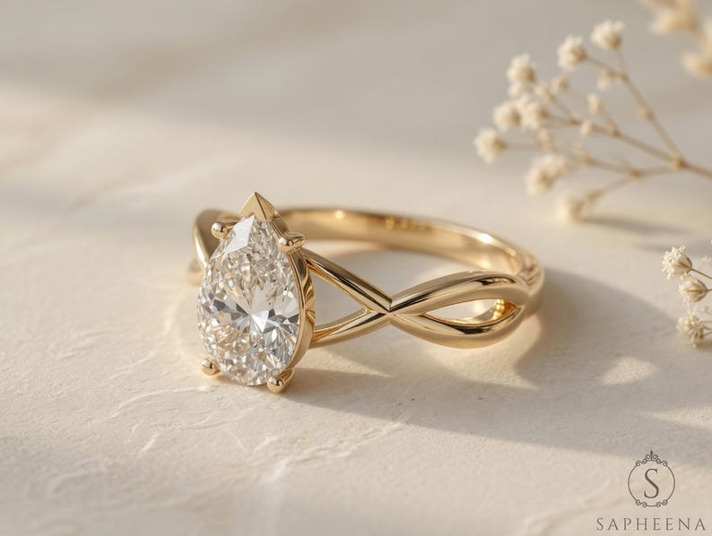 May include: A gold engagement ring with a pear-cut diamond. The ring has an infinity-style band. The diamond is the focal point, sparkling brightly. The ring is set against a neutral background, with delicate floral accents.