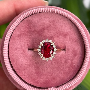 May include: A red oval gemstone ring with a halo of smaller white gemstones set in a gold band. The ring is displayed in a pink velvet ring box.