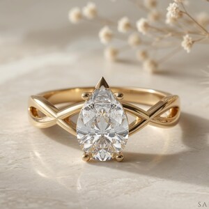 May include: A gold engagement ring with a pear-shaped diamond centre stone. The band features an infinity-style design. The ring is set on a light-coloured surface with soft lighting.