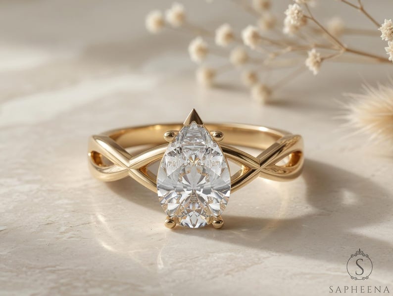 May include: A gold engagement ring with a pear-shaped diamond centre stone. The band features an infinity-style design. The ring is set on a light-coloured surface with soft lighting.