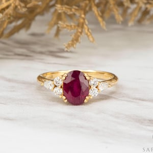 May include: A gold ring with a large oval ruby centre stone, complemented by smaller white gemstones. The ring's band has a textured pattern. The ring is set against a marble background with gold-coloured foliage.