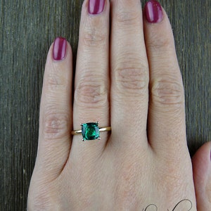 Op de afbeelding: Een gouden ring met een vierkante smaragdgroene edelsteen. De ring zit aan een vinger.