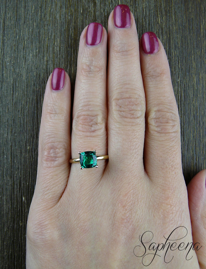 Op de afbeelding: Een gouden ring met een vierkante smaragdgroene edelsteen. De ring zit aan een vinger.