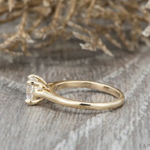 May include: A gold engagement ring with a round, clear center stone and a simple band. The ring is set on a wooden surface.