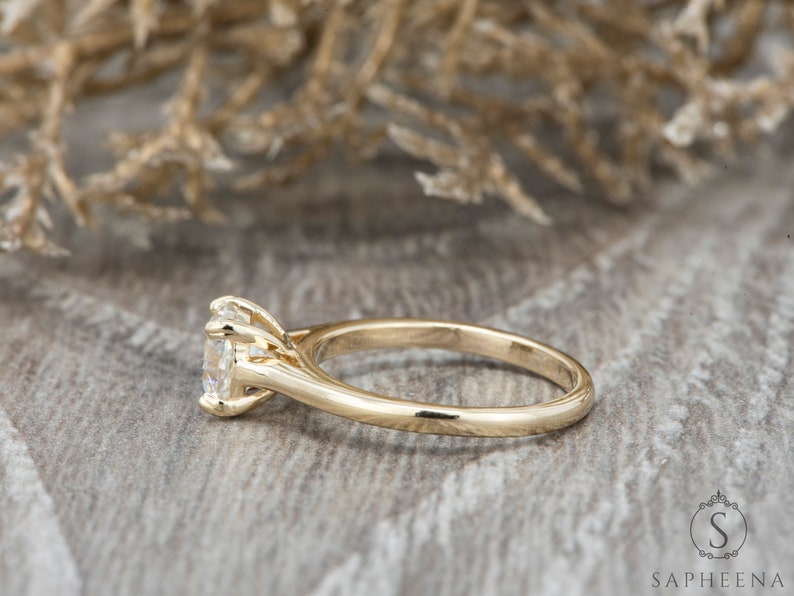 May include: A gold engagement ring with a round, clear center stone and a simple band. The ring is set on a wooden surface.