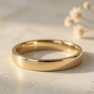 May include: A polished gold wedding band with a flat profile. The ring reflects light, and the background is a neutral colour with small white flowers. The ring is a simple, elegant design.