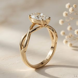 May include: A gold engagement ring with a pear-shaped diamond. The ring has an intricate, interwoven band design. The diamond is set in a bezel setting, and the ring is displayed on a light-coloured surface.