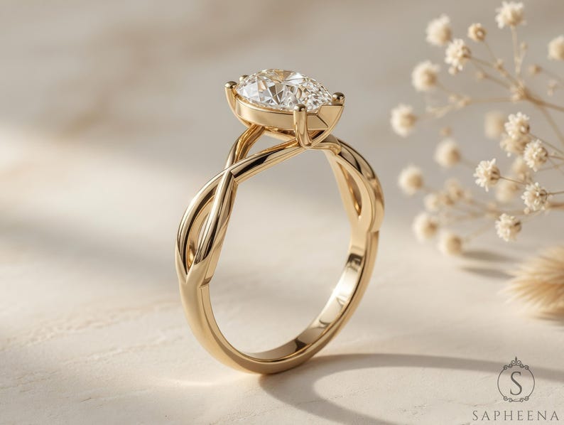May include: A gold engagement ring with a pear-shaped diamond. The ring has an intricate, interwoven band design. The diamond is set in a bezel setting, and the ring is displayed on a light-coloured surface.