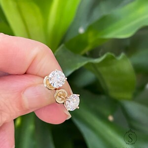 May include: A pair of gold stud earrings with clear round gemstones. The earrings are held in a hand against a green leafy background.