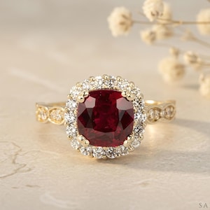 May include: A gold ring featuring a large, cushion-cut, deep red gemstone surrounded by a halo of smaller, clear gemstones. The band has a delicate, ornate design. The ring is set against a soft, neutral background, with a sprig of white flowers.