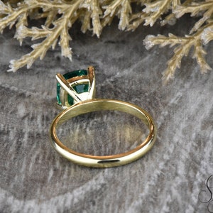 Op de afbeelding: Een gouden ring met een groene smaragd gezet in een kraalbezetting. De ring ligt op een grijs, getextureerd oppervlak.