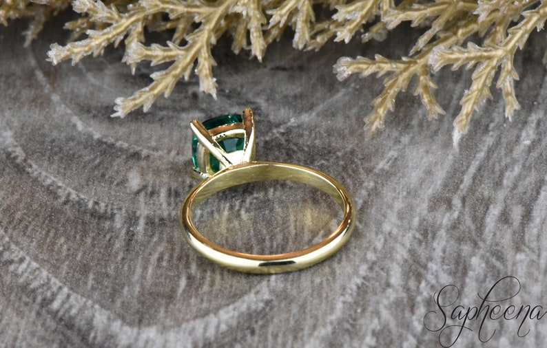 Op de afbeelding: Een gouden ring met een groene smaragd gezet in een kraalbezetting. De ring ligt op een grijs, getextureerd oppervlak.