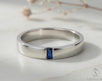May include: A silver ring featuring a rectangular, dark blue gemstone embedded in the band. The ring has a flat, polished surface and a simple design. This is a piece of jewellery.
