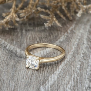May include: A gold engagement ring with a round, clear center stone. The ring is set on a wooden surface.