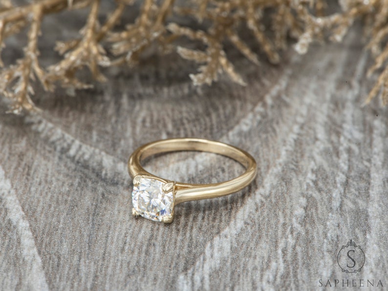 May include: A gold engagement ring with a round, clear center stone. The ring is set on a wooden surface.