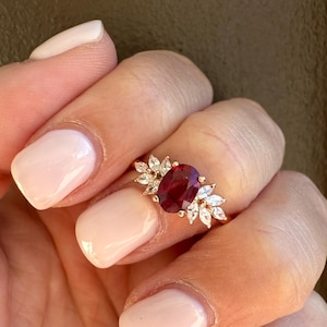 May include: A gold ring with a large oval ruby gemstone at its centre, surrounded by smaller white gemstones. The ring is set on a gold band and is shown on a hand with light pink nail polish.