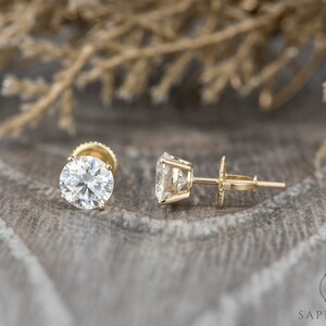 May include: A pair of gold stud earrings with round, clear gemstones. The earrings are set against a brown, textured background.