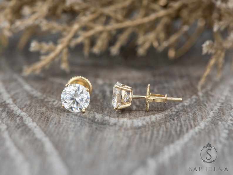 May include: A pair of gold stud earrings with round, clear gemstones. The earrings are set against a brown, textured background.