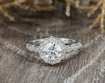 May include: A silver engagement ring with a round, clear center stone surrounded by smaller stones in a floral halo setting. The band is decorated with a delicate, scalloped design.