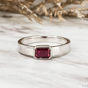 May include: A silver ring with a rectangular, deep red gemstone. The ring has a simple, polished band and a bezel setting for the gemstone. The ring is displayed on a white marble surface.
