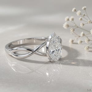 May include: A silver engagement ring with a pear-cut diamond. The ring has an infinity-style band design. The diamond is the focal point, reflecting light. The ring is set against a soft, neutral background, with a sprig of white flowers.