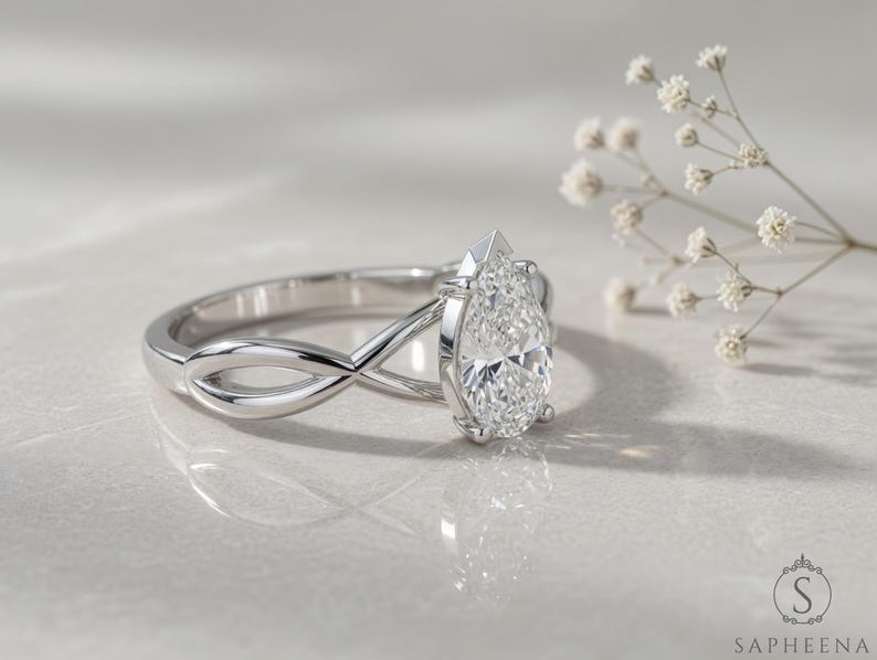 May include: A silver engagement ring with a pear-cut diamond. The ring has an infinity-style band design. The diamond is the focal point, reflecting light. The ring is set against a soft, neutral background, with a sprig of white flowers.