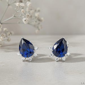 Puede incluir: Un par de pendientes de botón con una gran piedra azul zafiro en forma de pera, rodeada por un halo parcial de pequeñas piedras blancas redondas y transparentes sobre una base de metal plateado. El logotipo SAPHEENA es visible.