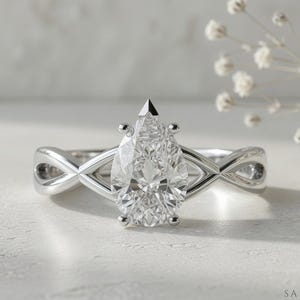 May include: A silver engagement ring with a pear-cut diamond. The band has an infinity design. The ring is set against a soft, neutral background with delicate white flowers.