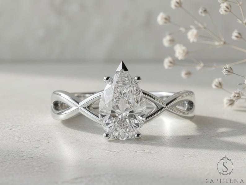 May include: A silver engagement ring with a pear-cut diamond. The band has an infinity design. The ring is set against a soft, neutral background with delicate white flowers.