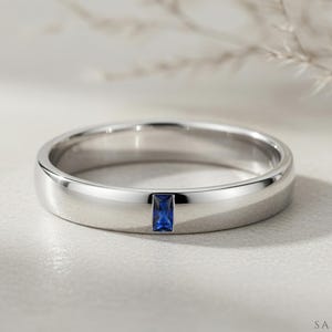 Puede incluir: Un anillo de banda en tono plateado pulido que presenta una única piedra preciosa de zafiro azul de corte baguette rectangular, engastada verticalmente al ras en la superficie metálica lisa y redondeada del anillo.