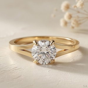 May include: A gold engagement ring with a large, round, clear gemstone. The ring has a split shank design, with the band splitting on either side of the gemstone. The ring is set against a neutral background with soft lighting.