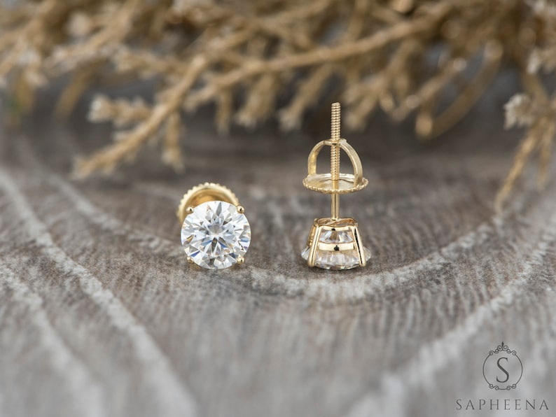 May include: A pair of gold stud earrings with a round, clear gemstone in the center. The earrings have a screw-back closure.