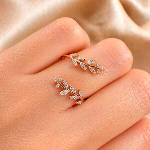 May include: Two gold-colored rings, each featuring a delicate leaf design adorned with small, sparkling gemstones. The rings are displayed on a hand, showcasing their intricate details and elegant style. The rings are a beautiful piece of jewelry.