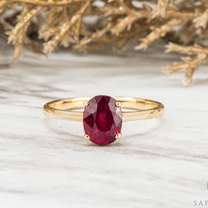Könnte beinhalten: Ein goldener Ring mit einem großen, oval geschliffenen Rubin-Edelstein. Der Rubin hat eine tiefrote Farbe und ist der Mittelpunkt des Rings. Der Ring liegt auf einer Marmoroberfläche mit verschwommenem Hintergrund.
