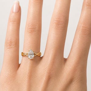 May include: A gold ring with a pear-cut diamond centre stone. The band has an interwoven design. The ring is shown on a person's hand, highlighting the ring's intricate details and sparkle.